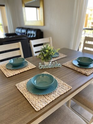 Uma mesa de jantar com quatro tigelas azul-turquesa e pratos combinando, colocados sobre jogos americanos de palha. Uma pequena planta em vaso está posicionada ao centro.