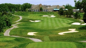 Een golfbaan met bunkers, een slingerende weg en een clubhuis op de achtergrond.