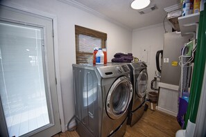 Une buanderie équipée de deux lave-linge et deux sèche-linge à chargement frontal, d’une fenêtre avec des stores et d’une étagère contenant des produits de nettoyage.