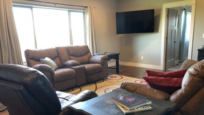 Una sala de estar con un sofá de cuero marrón, un sillón reclinable, una mesa de centro con libros, un televisor de pantalla plana montado en la pared y una puerta que da a otra habitación.