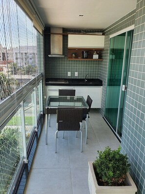 Un balcón con una mesa y sillas de vidrio, una pequeña zona de cocina con un lavabo y armarios, y vistas a la ciudad.