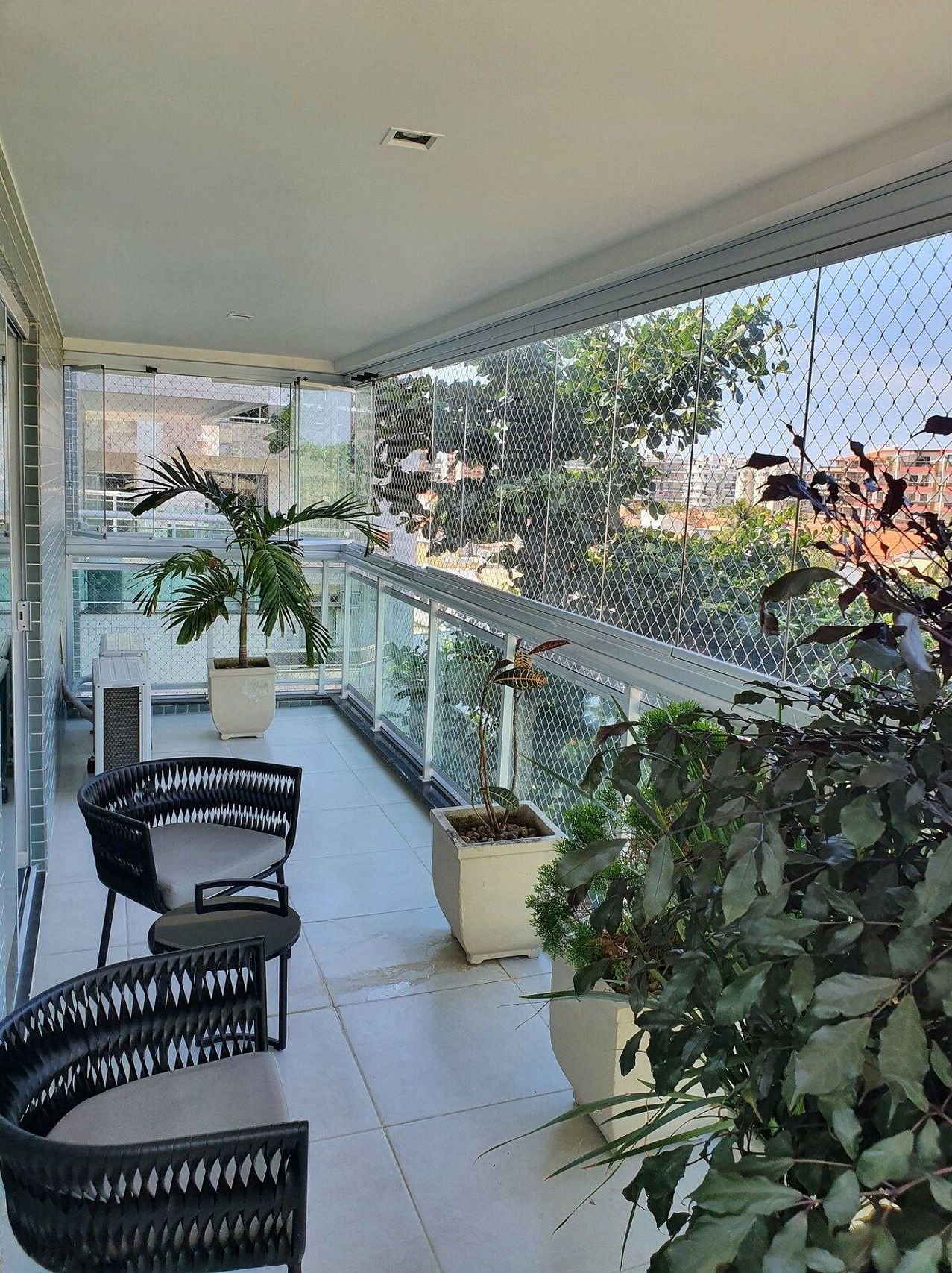 Uma varanda com cadeira, plantas em vasos e uma grade de arame que dá para uma vista urbana.