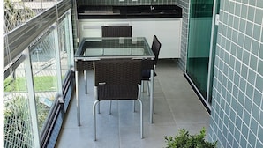A small balcony with a glass table and chairs, a potted plant, and a kitchen area with green tiles and cabinets. - Lindo AP completo no Braga em Cabo Frio (Cabo Frio)