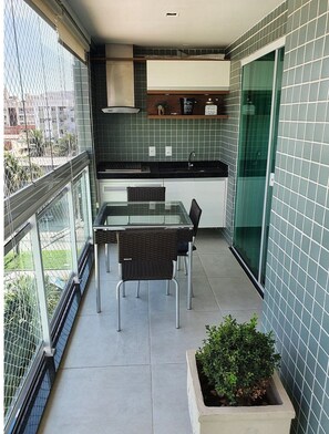 A small balcony with a glass table and chairs, a potted plant, and a kitchen area with green tiles and cabinets.