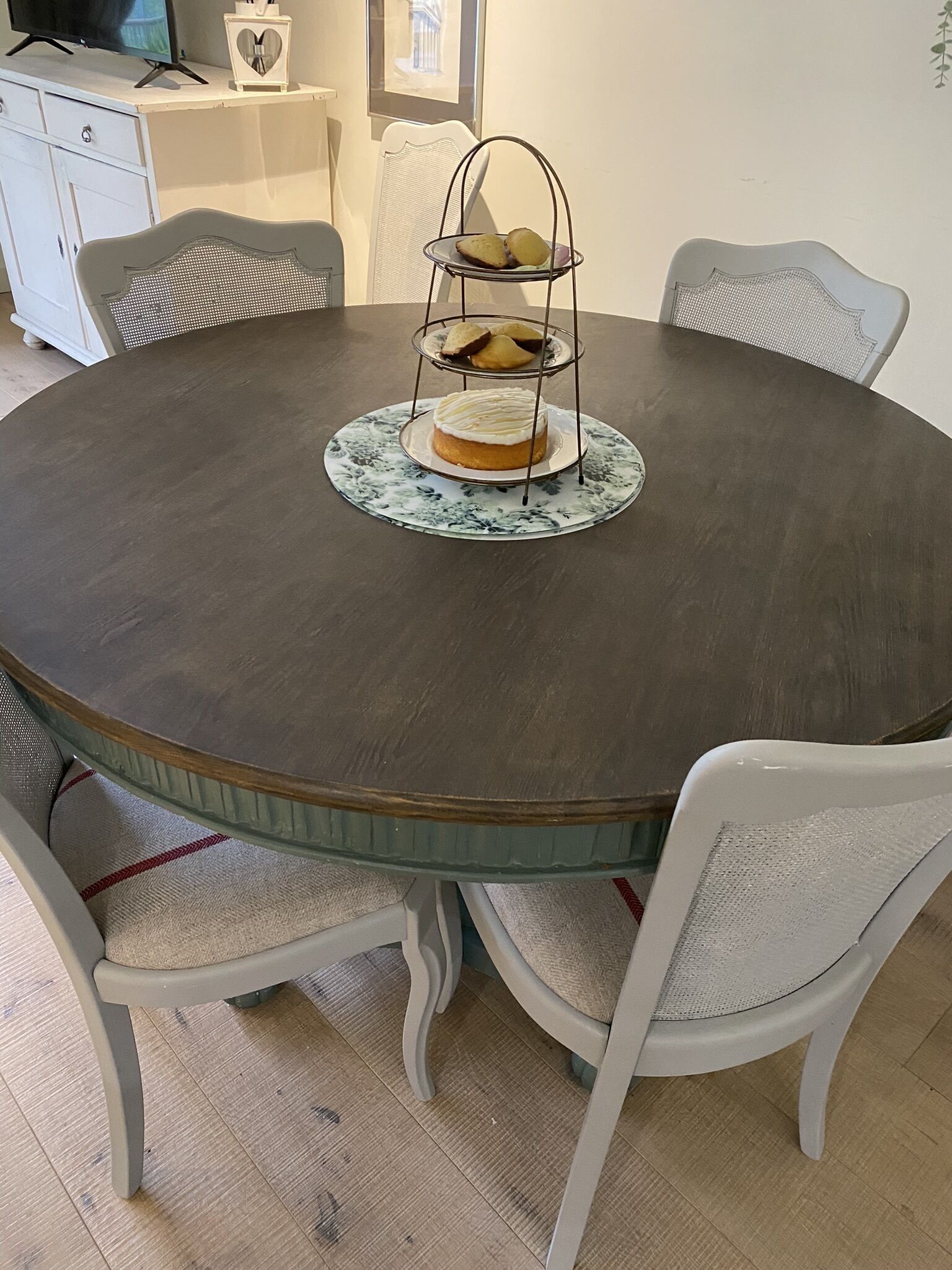 A round dining table with a dark wooden top, surrounded by white chairs with woven seats. A three-tiered cake stand with a cake on the top tier is placed on a patterned placemat.