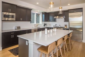 Cocina moderna con armarios oscuros, electrodomésticos de acero inoxidable, una isla grande con taburetes altos e iluminación colgante.
