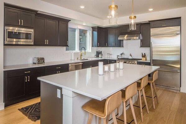 A modern kitchen with dark cabinets, stainless steel appliances, a large island with bar stools, and pendant lighting.