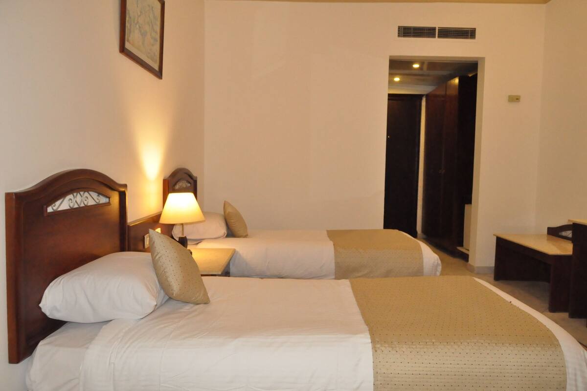 Habitación de hotel con dos camas, una mesita de noche con lámpara, un banco de madera y una puerta que da a otra habitación.