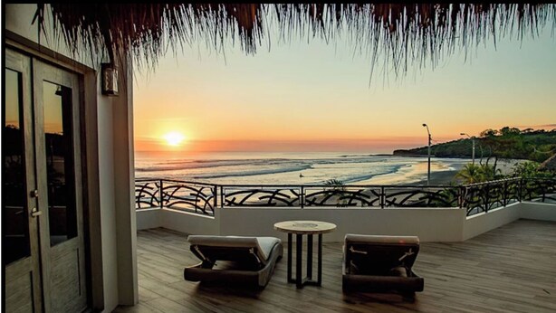 Een balkon met houten vloer, twee lounge fauteuils, een klein rond tafeltje en uitzicht op de oceaan bij zonsondergang.