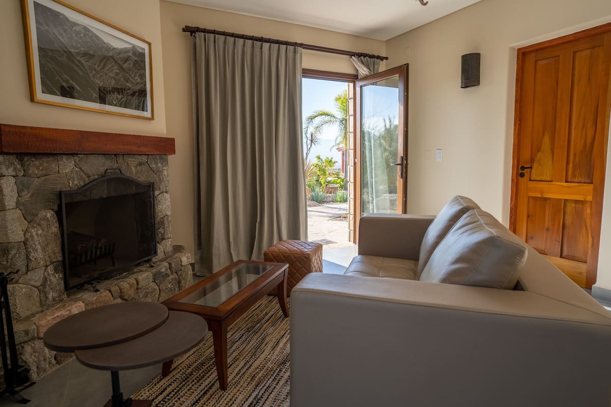Ein Wohnzimmer mit Kamin aus Stein, einer Couch, einem Couchtisch und Blick auf einen Garten durch eine Glastür.