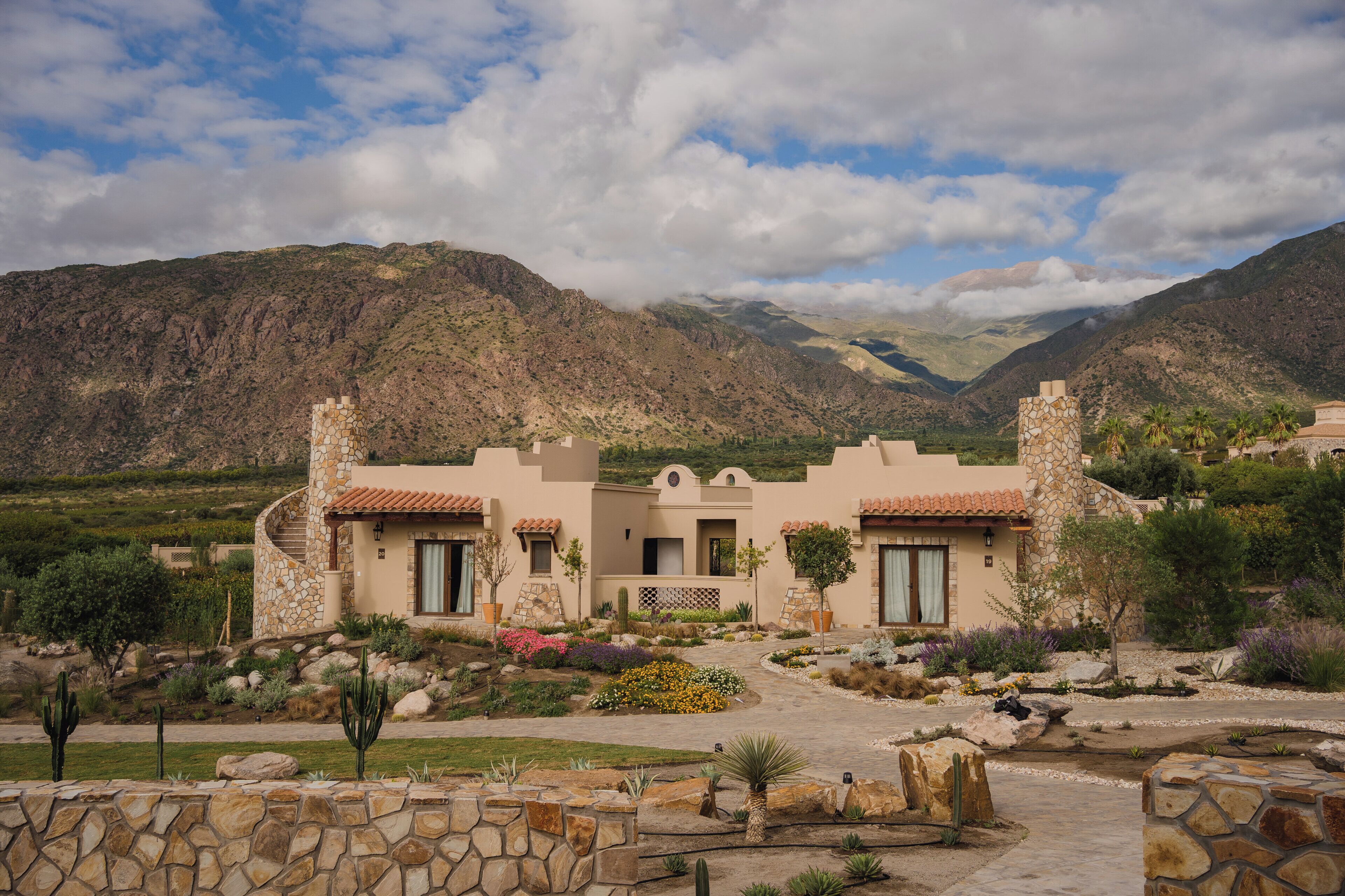 A Mediterranean-style villa with a stone facade, multiple windows, and a landscaped garden with cacti and flowering plants.
