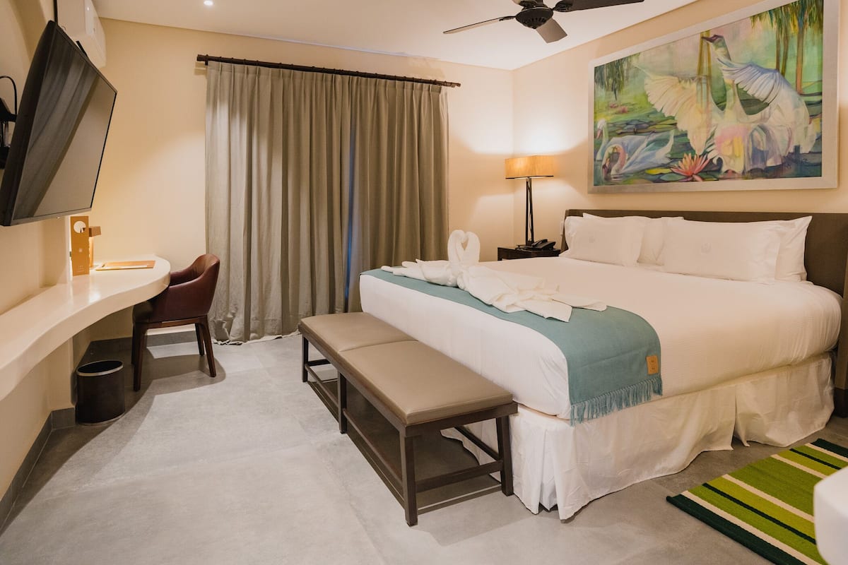 Ein Hotelzimmer mit Bett, Schreibtisch, Stuhl, Bank, einem Gemälde und einem Deckenventilator.