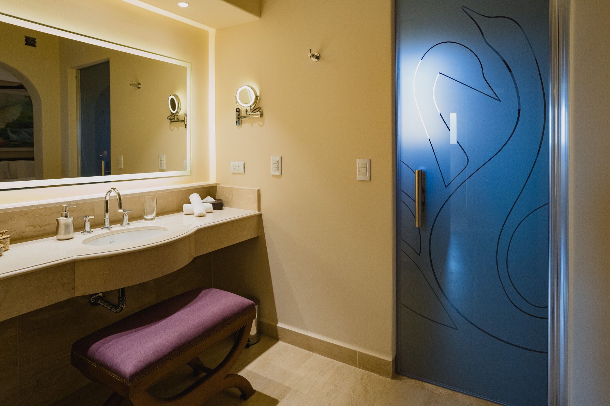 Een badkamer met een grote spiegel, twee wastafels, een bankje en een deur met een bijzonder ontwerp.