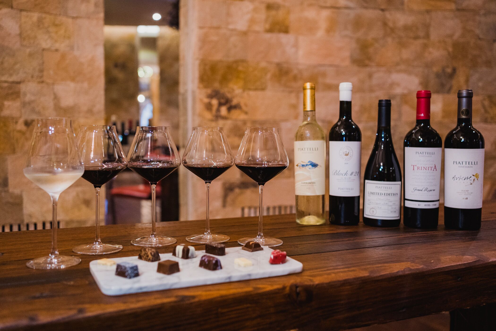 A wooden table with wine glasses and bottles of wine, a marble serving tray with chocolates, and a stone wall background.