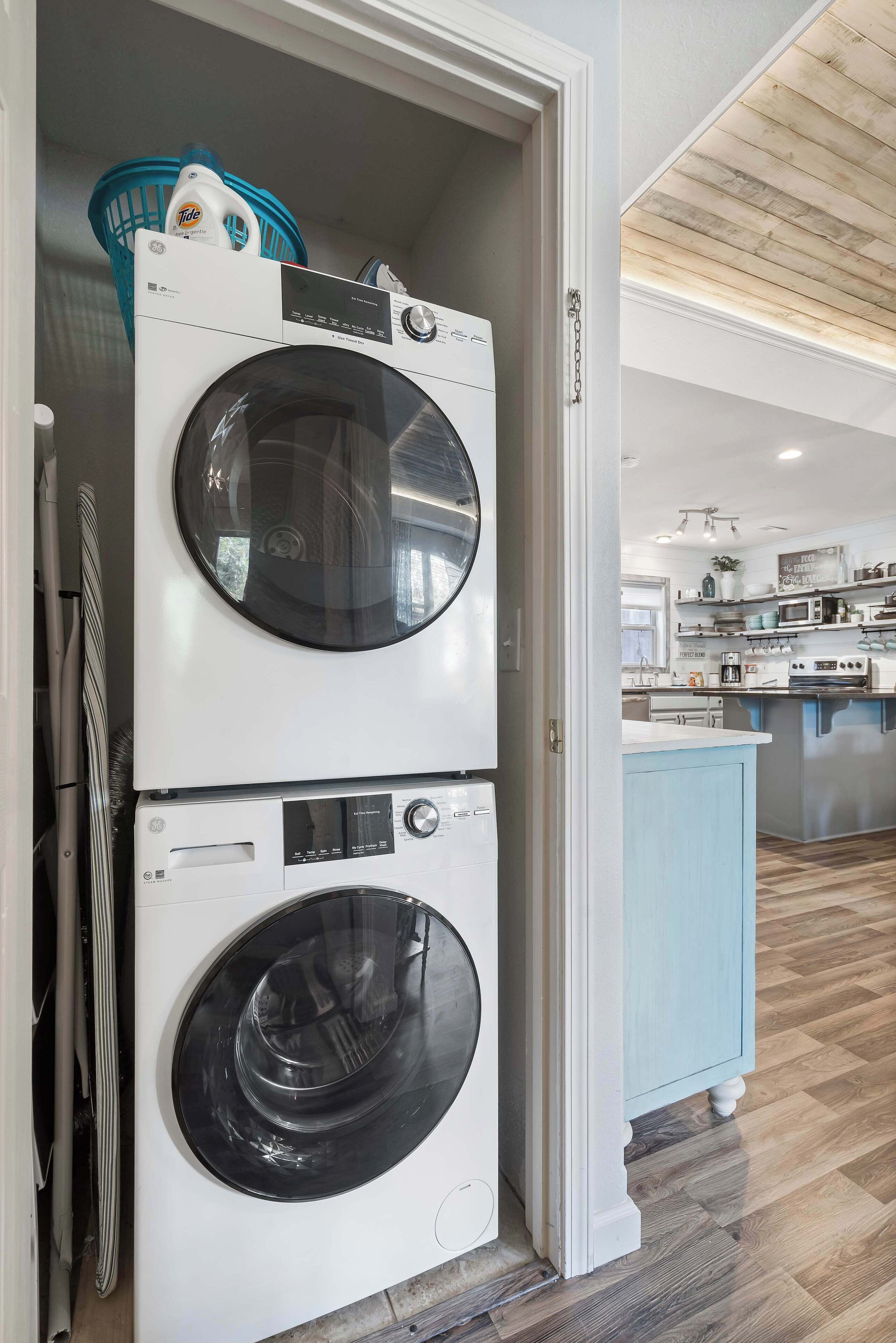 Un cuarto de lavado con dos lavadoras de carga frontal apiladas, una cesta de ropa azul y una zona de cocina al fondo.