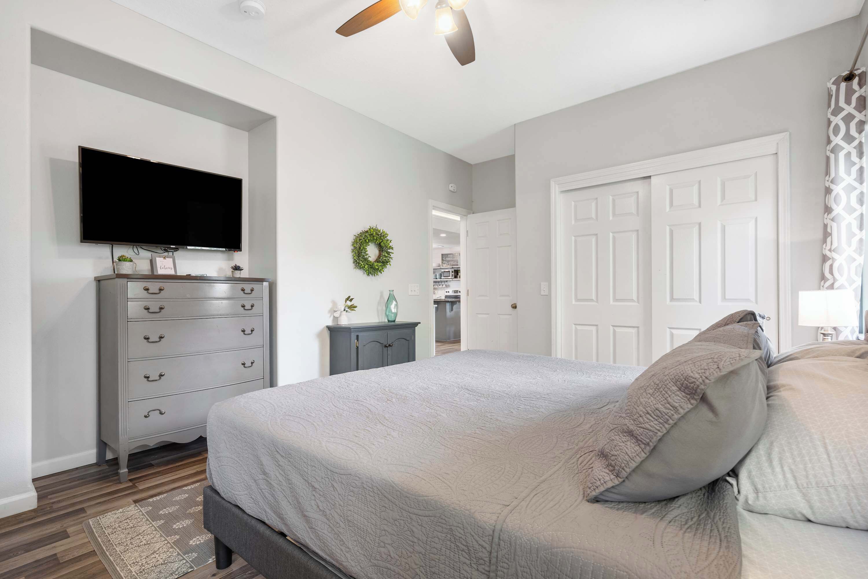 Une chambre à coucher comprenant un lit, une commode, un téléviseur, un ventilateur de plafond et une porte donnant sur une autre pièce.