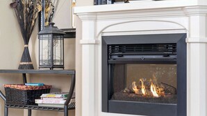 Une cheminée avec un feu de bois, une lanterne noire et un panier en osier rempli de livres, le tout posé sur une étagère en métal.