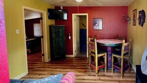 A living room with a wooden floor, a sofa with a pink and blue blanket, a dining table with chairs, a television mounted on the wall, and a red wall with a deer skull decoration. - Cozy cabin at Inn at Lake Roberts (Lake Roberts)