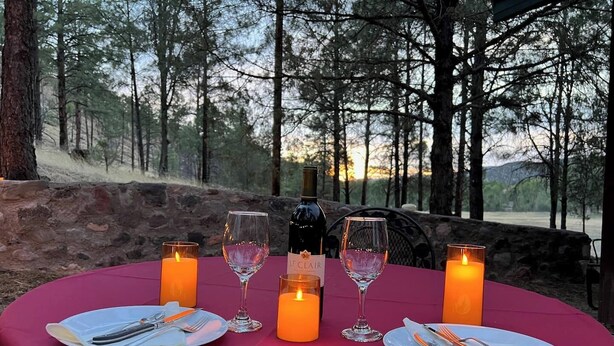 Mesa para dos personas dispuesta con velas, vino y cubiertos, rodeada de un paisaje boscoso.