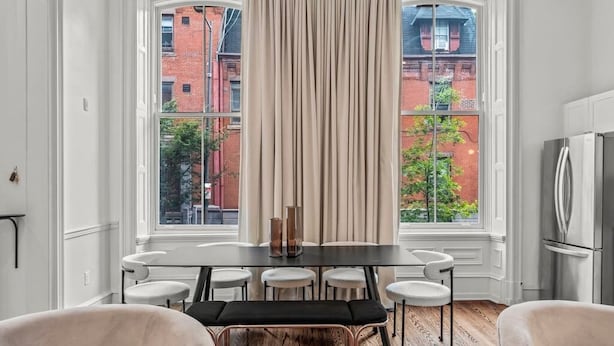 Ein Essbereich mit Tisch, Stühlen und einer Bank. Große Fenster mit Vorhängen, ein Kühlschrank und Blick auf Gebäude.