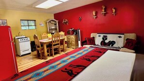 Una habitación compacta con cama, un rincón comedor con mobiliario de madera, una pared roja y una pequeña kitchenette con estufa.