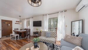 Ein geräumiges Wohnzimmer mit Holzboden, einem grauen Sofa, einem Couchtisch mit Pflanze, einem an der Wand montierten Fernseher und einem Essbereich mit Holztisch und Stühlen.