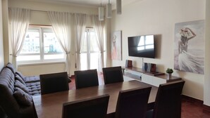 Ein modernes Wohnzimmer mit Sofa, Esstisch, Stühlen, einem Flachbildfernseher, Kunstwerken und einem fenster mit Vorhängen.
