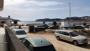 Ein Parkplatz mit mehreren Autos, ein Gebäude mit einem Schild und im Hintergrund Meer und Hügel.