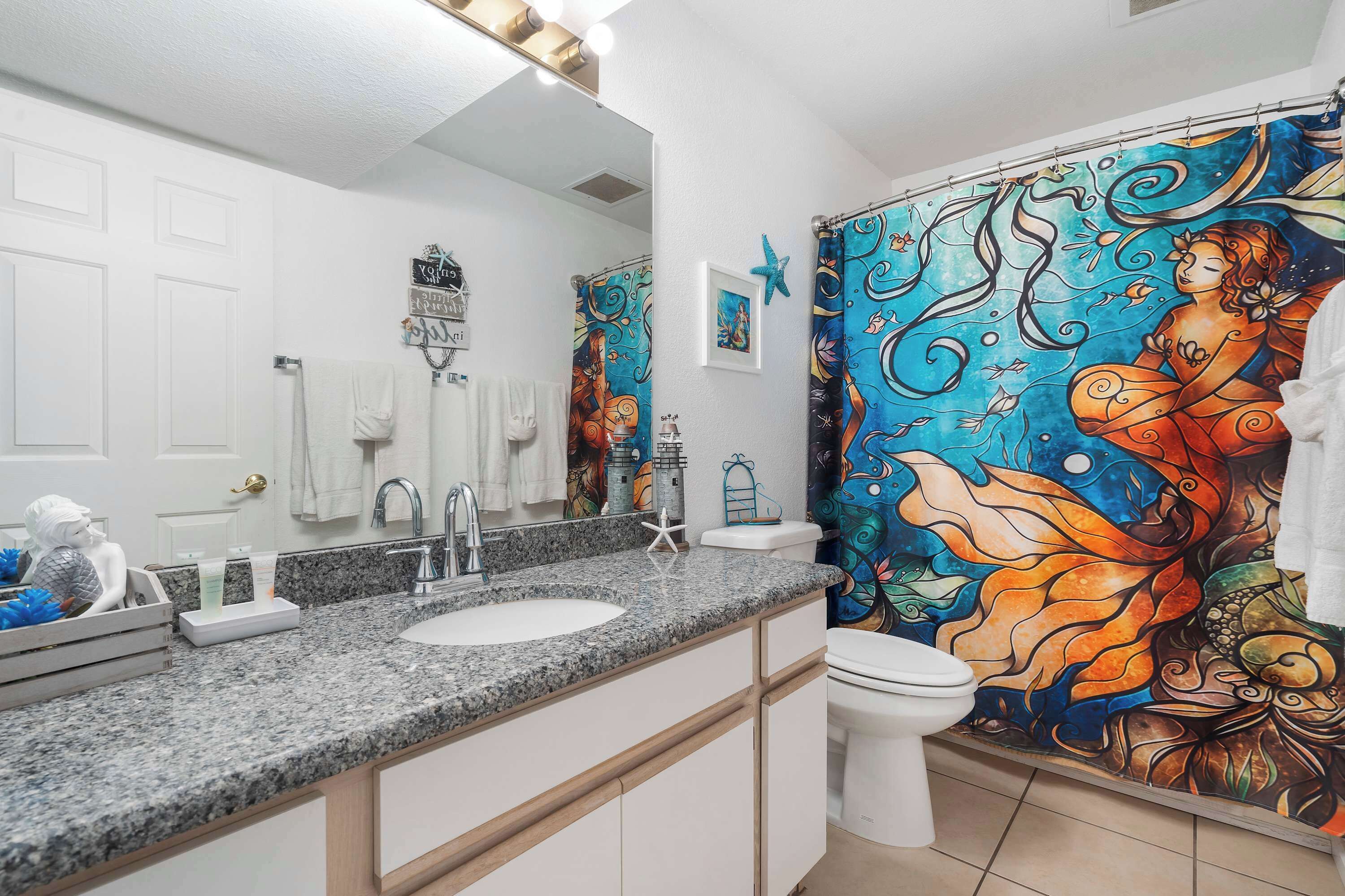A bathroom with a large mirror, a granite countertop, a white sink, a toilet, and a colorful shower curtain.