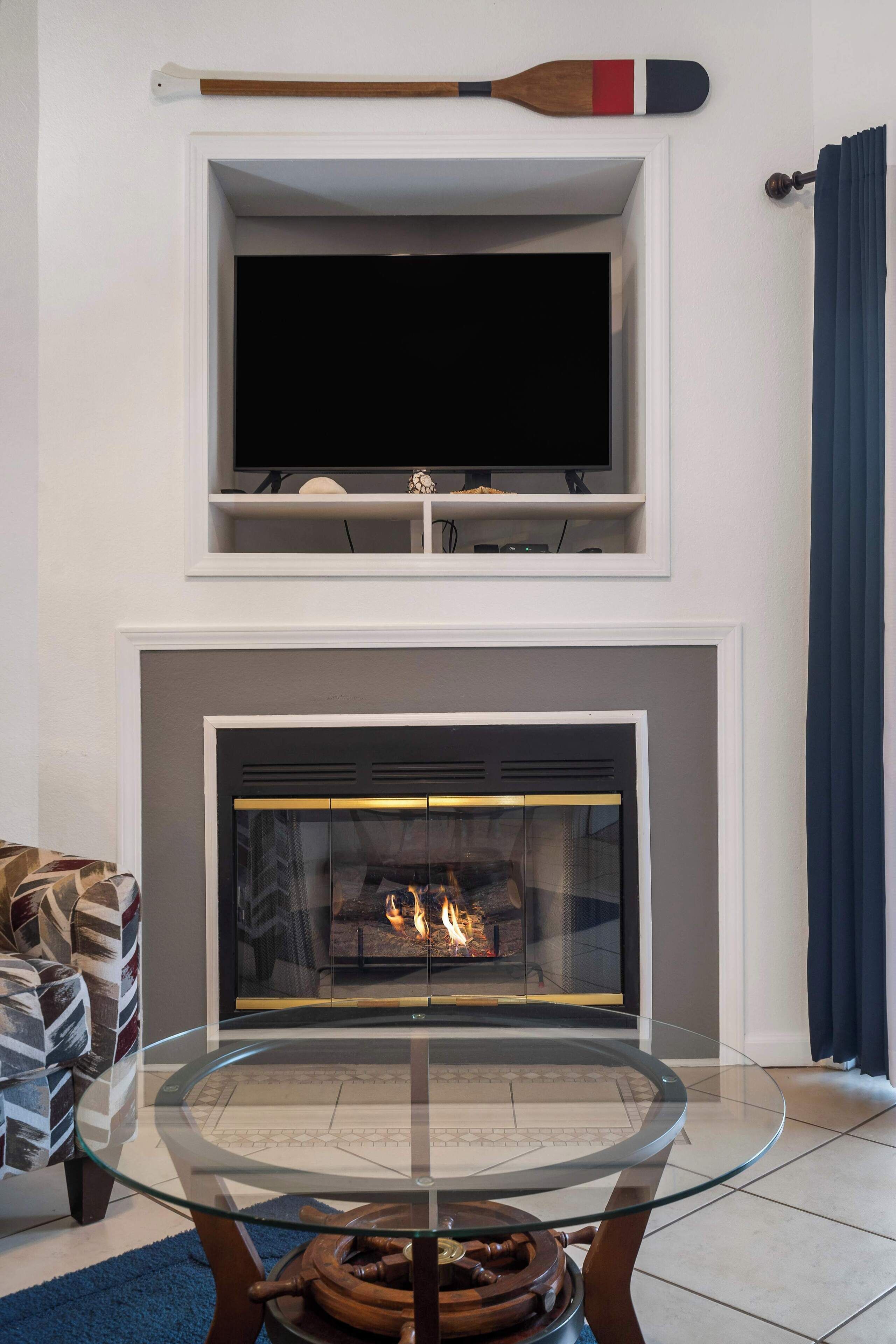 A living room with a fireplace, a glass-top coffee table, and a television above the fireplace.