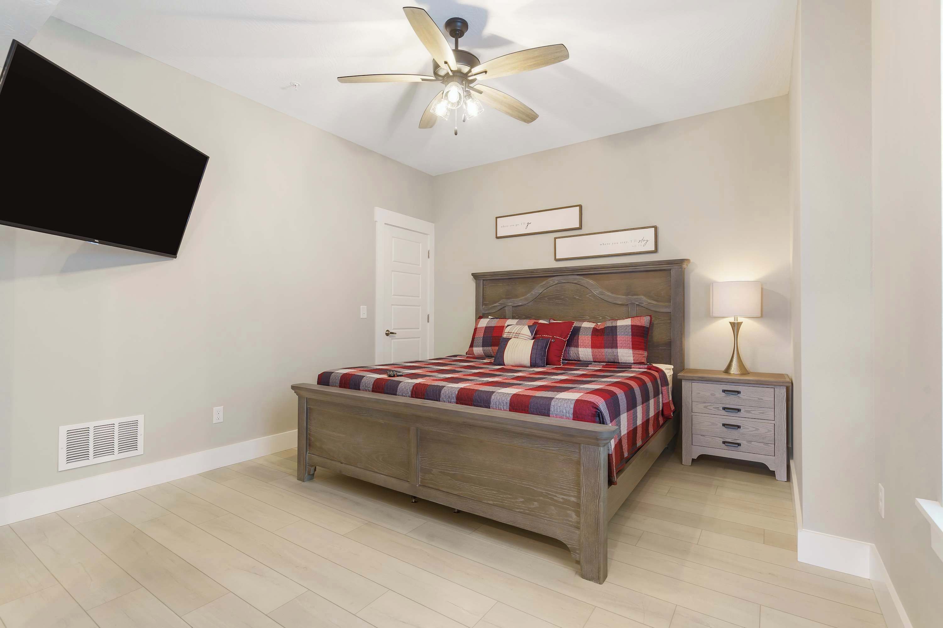 A bedroom with a bed, a nightstand, a wall-mounted TV, a ceiling fan, and a door.