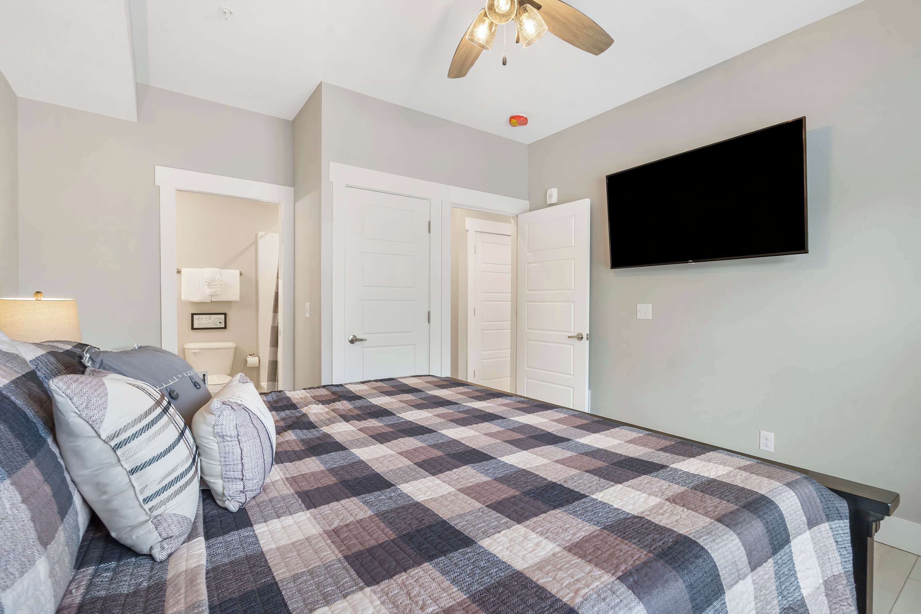 Une chambre à coucher avec une couvre-lit à carreaux, un ventilateur de plafond, un téléviseur à écran plat et des portes blanches.