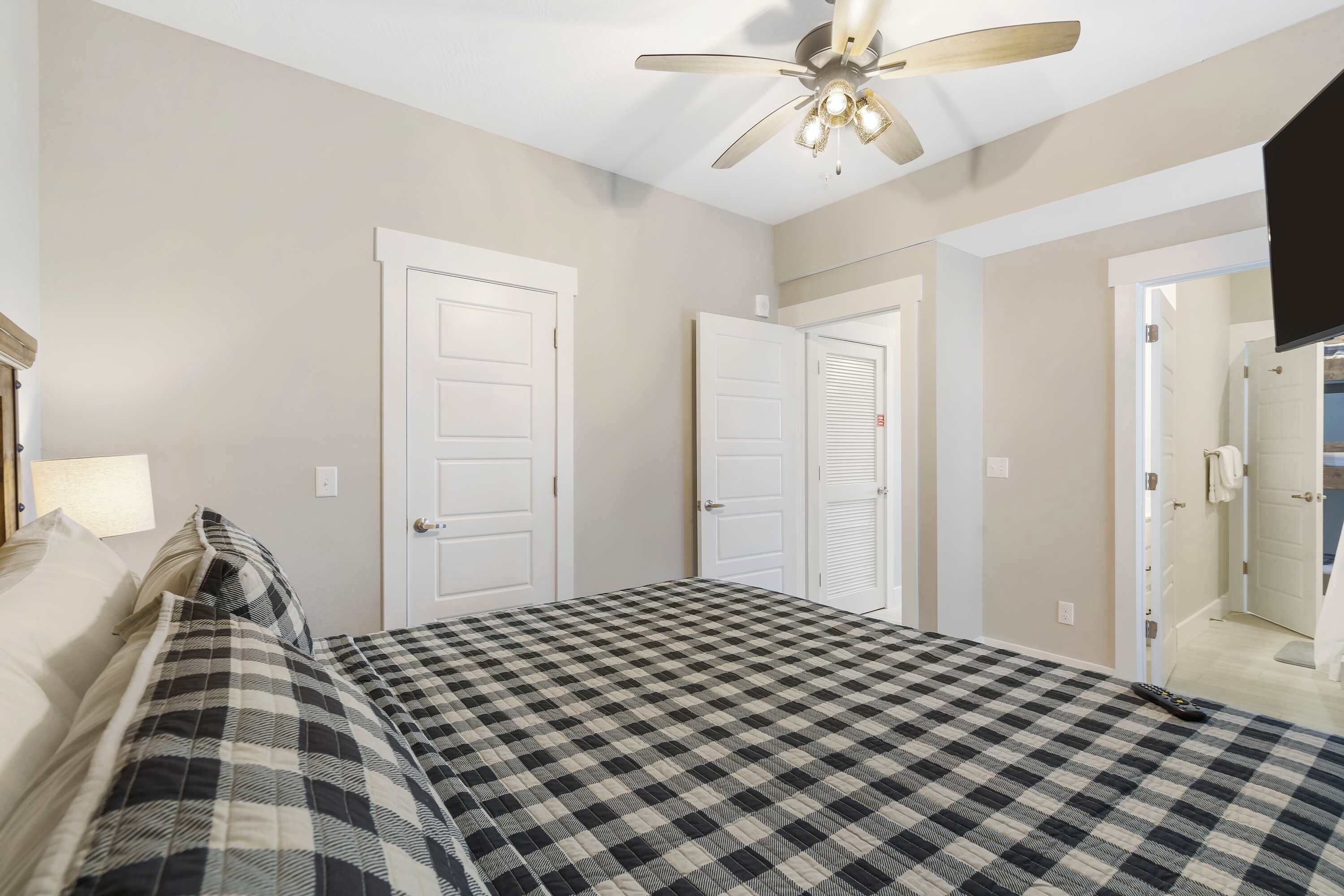 A bedroom with a checkered bedspread, white doors, a ceiling fan, and a TV mounted on the wall.