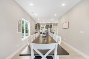A dining room with a long table, white chairs, a potted plant, and a kitchen in the background. - Hilltop Haven at Table Rock - Lake Views & Nearby Bransons Best Attractions (Branson)