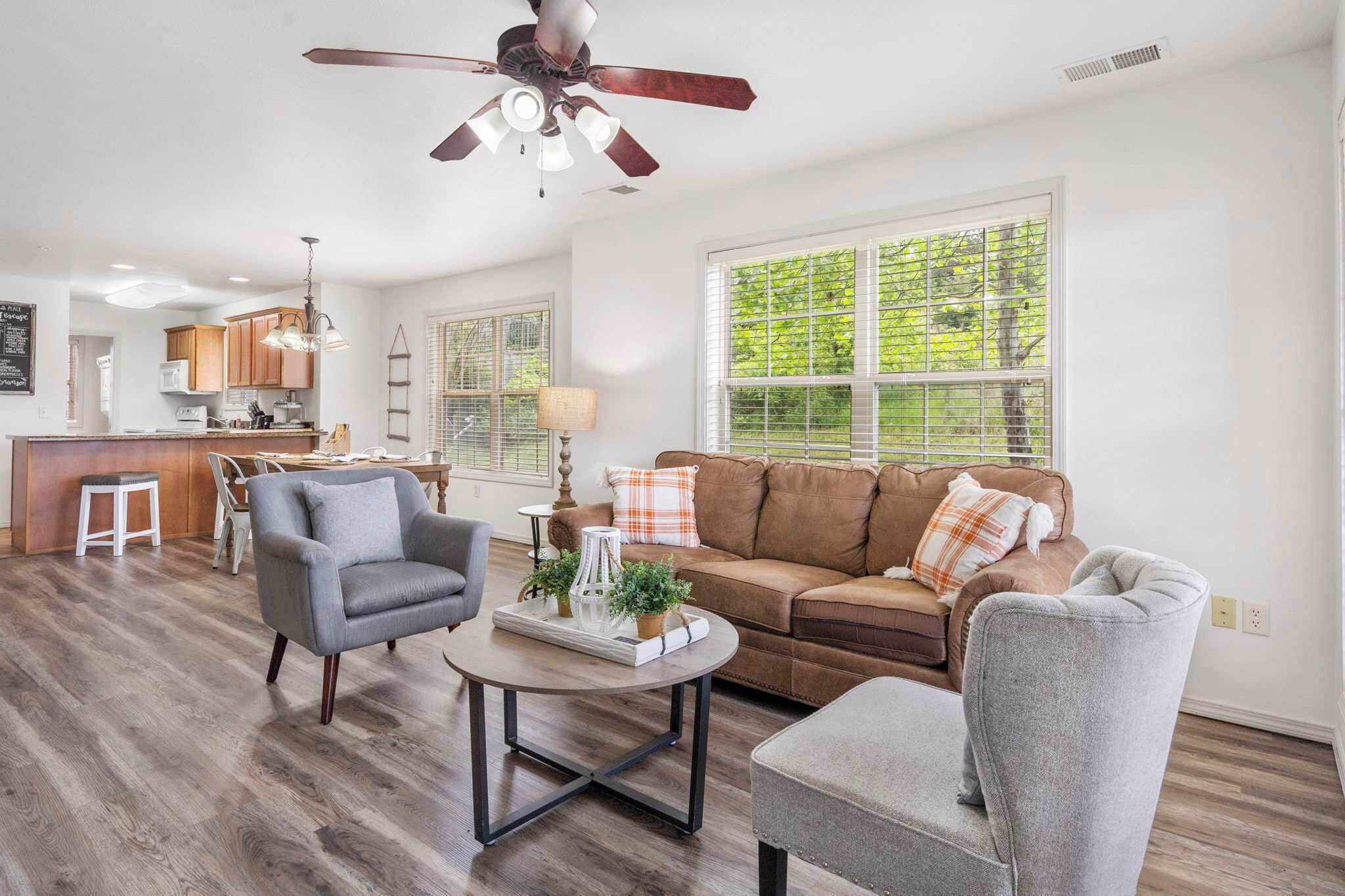 A spacious living room with a brown leather sofa, grey armchairs, and a coffee table. A ceiling fan and pendant lights are visible.