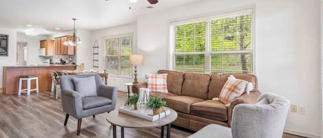 A spacious living room with a brown leather sofa, grey armchairs, and a coffee table. A ceiling fan and pendant lights are visible.