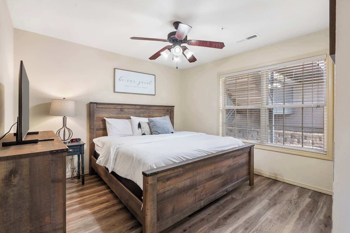 A bedroom with a wooden bed, a ceiling fan, a bedside table with a lamp, and a window with blinds.