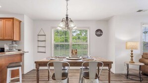 A dining area with a wooden table, white chairs, a chandelier, and a kitchen in the background. - Retreat at Par Lane - Family Friendly Condo (Branson West)