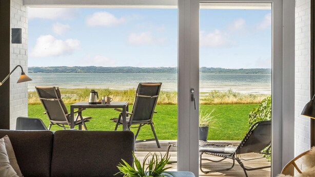 A modern room with a large sliding glass door leading to a deck overlooking a lake. There are two lounge chairs and a small table with a coffee pot and mug.