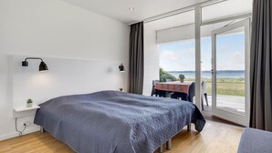 A modern bedroom with a large bed, a small table with a tablecloth, and a view of the outdoors through a sliding glass door.