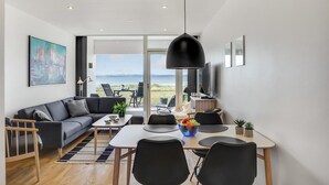 A modern living room with a sofa, coffee table, and dining area. There is a large window with a view of the outdoors.