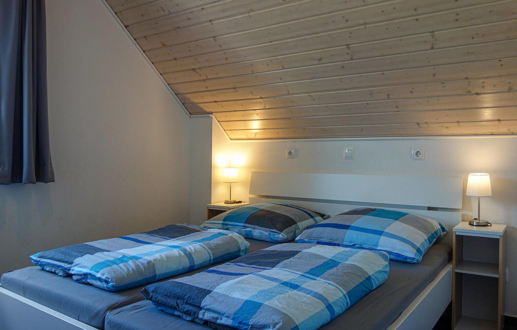 Una habitación con techo inclinado, dos camas con ropa de cama a cuadros azul y blanco, lámparas de noche y una mesita de noche de madera.