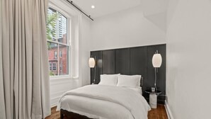 A modern bedroom with a large bed, two bedside lamps, a window with curtains, and a view of buildings outside. - Luxury 1BD Rittenhouse Sq. | Hosted By Stay Rafa (Philadelphia)