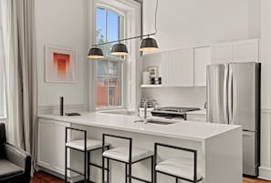 Une cuisine moderne avec des meubles blancs, un îlot central doté de tabourets de bar, un réfrigérateur en acier inoxydable et des luminaires suspendus.