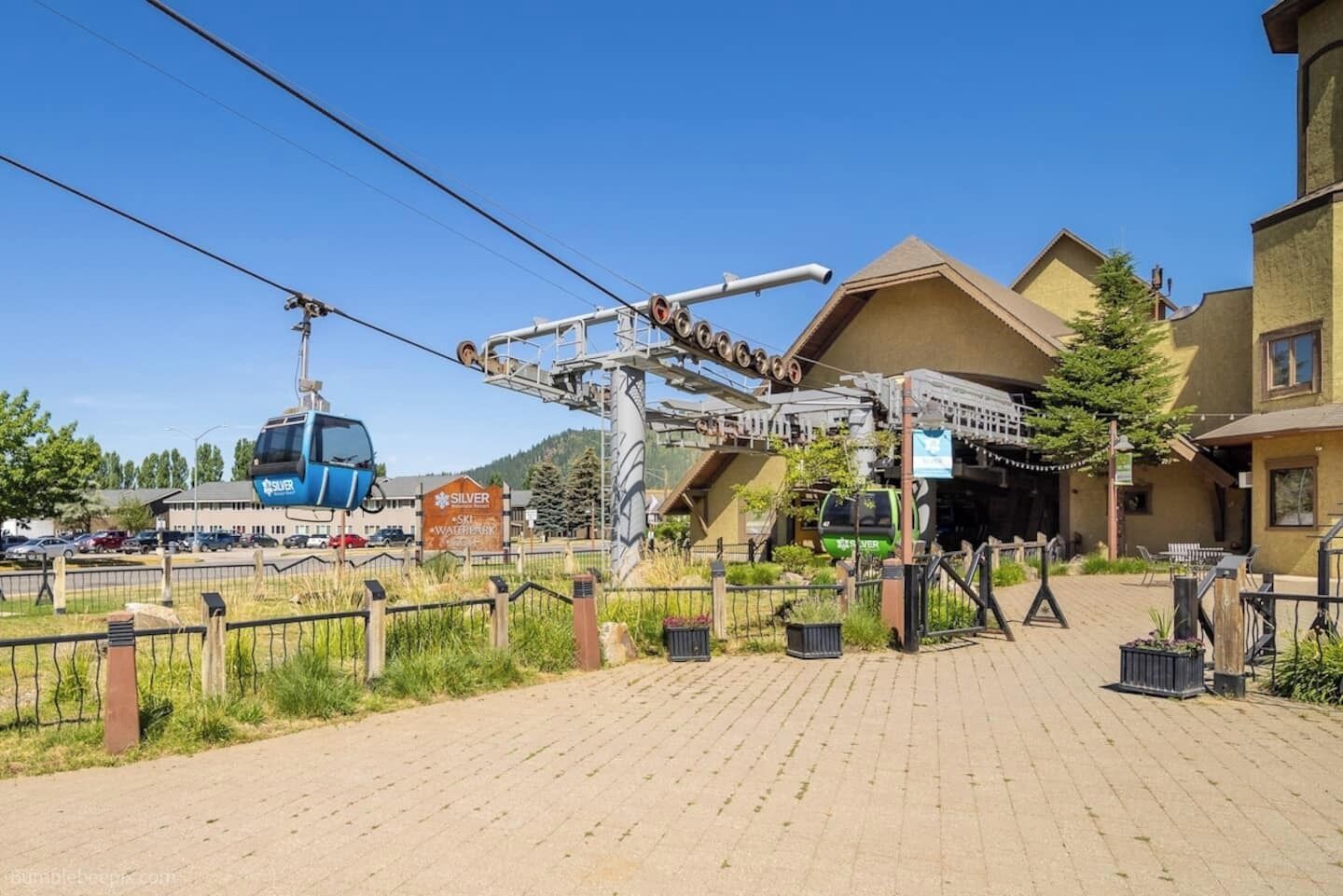 A ski lift station with a cable car and a gondola, a paved area with a fence, and buildings in the background.