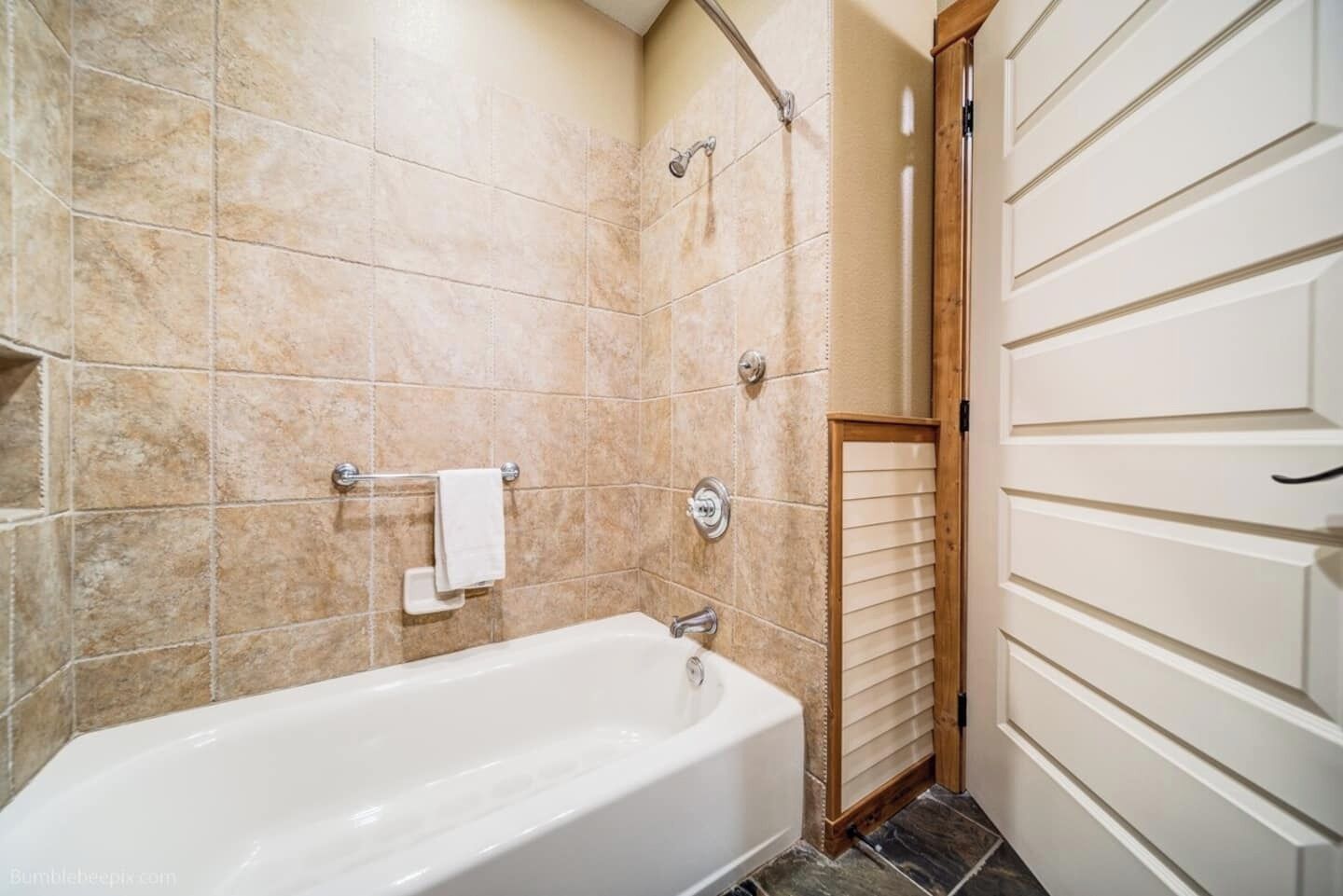 A bathroom with a bathtub, beige tiles, a towel rack, and a wooden door.