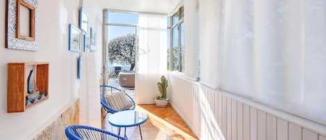 Ein schmaler Gang mit blauen Stühlen und einem Tisch, der zu einem Außenbereich mit Meerblick führt.