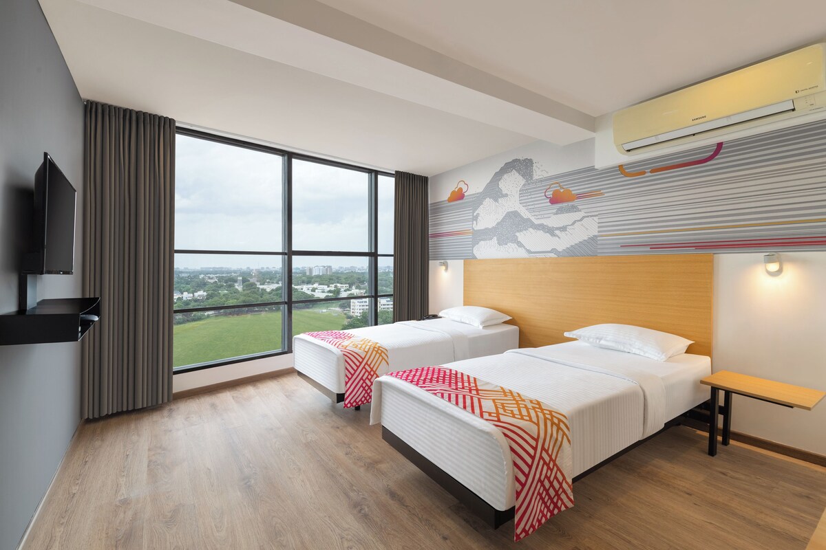 Habitación de hotel con dos camas, un ventanal grande con vistas a la ciudad, un equipo de aire acondicionado y un televisor montado.