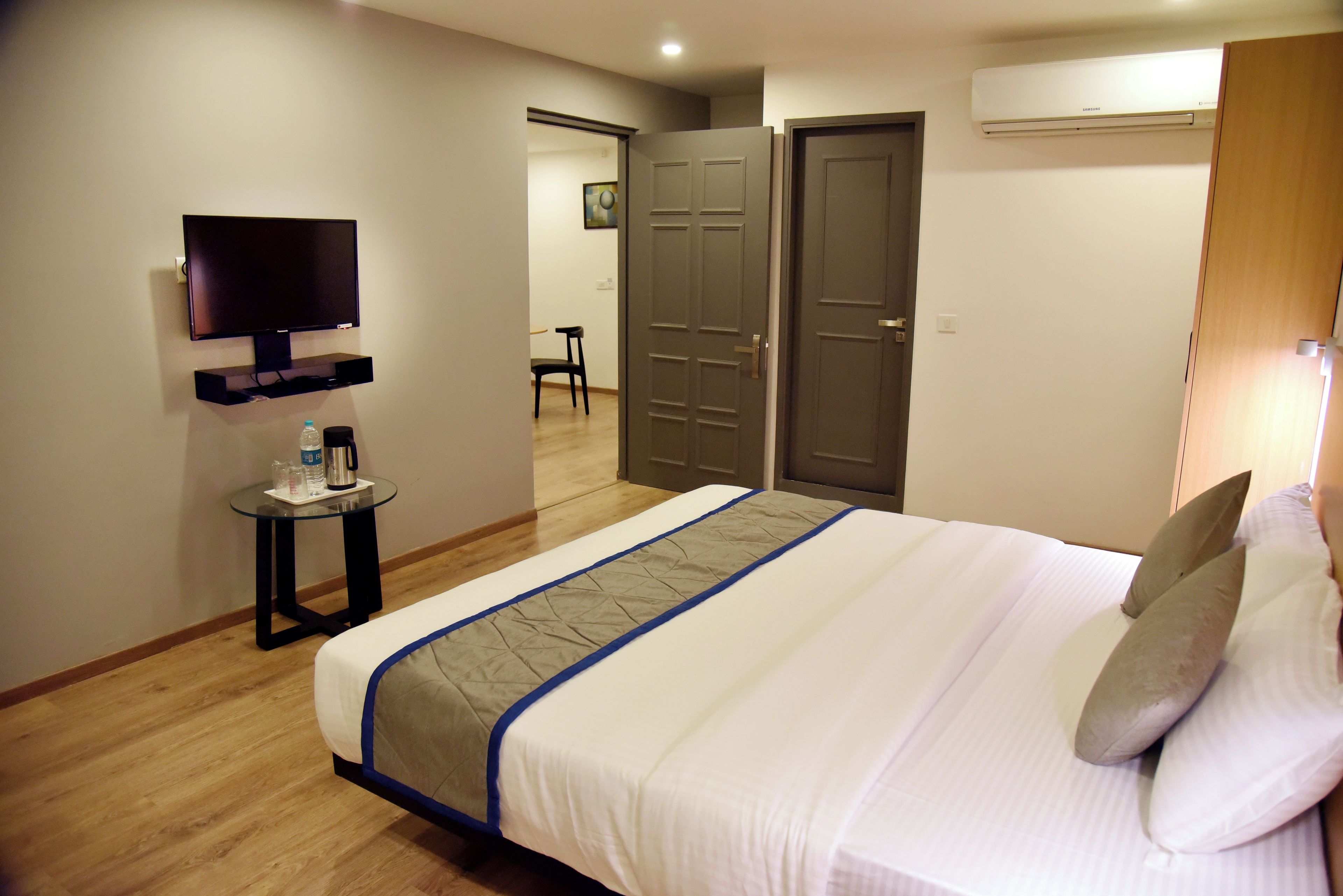 A hotel room with a bed, a television mounted on the wall, a small table with a bottle and a cup, and a door leading to another room.