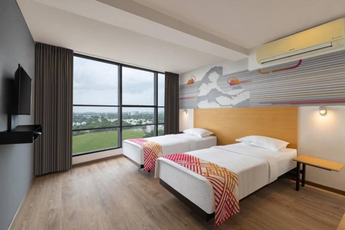 A hotel room with two beds, a large window with a city view, a mounted TV, and an air conditioning unit.